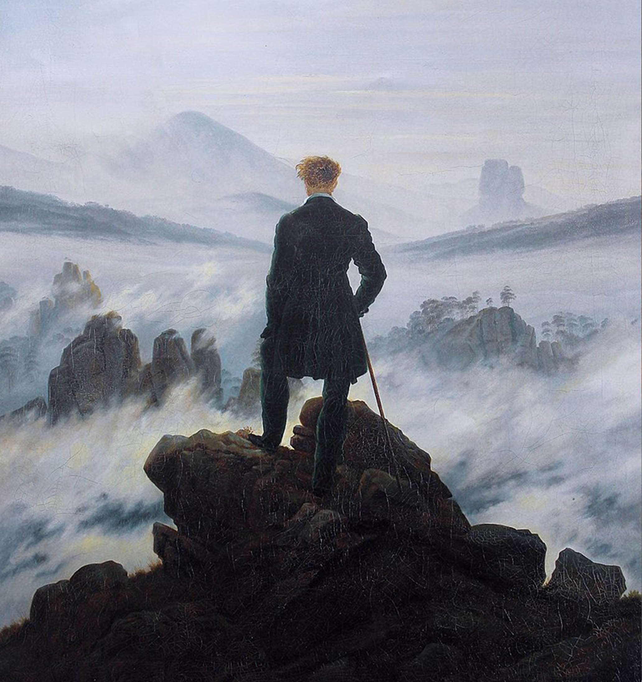 Man in black with walking cane stands upon a craggy height just above the fog. Looking away from us, he gazes upon the seemingly endless mist-shrouded landscape before him.